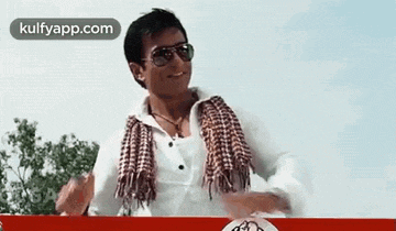 a man wearing sunglasses , a scarf and a white shirt is dancing .