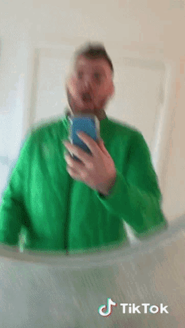 a man in a green jacket is taking a selfie in front of a mirror with tiktok written on the bottom right