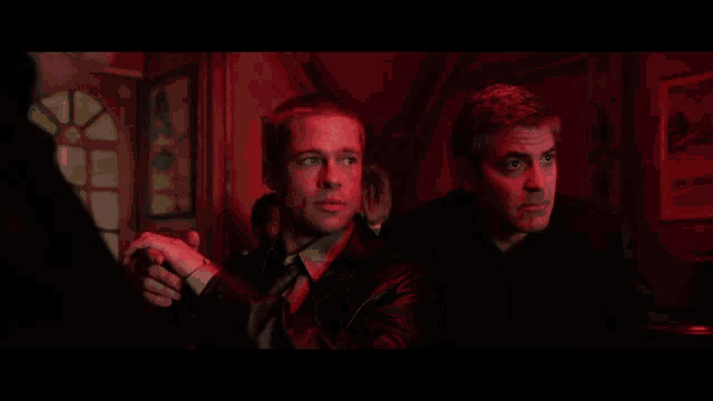 two men are sitting in a dark room and one of them is wearing a black jacket