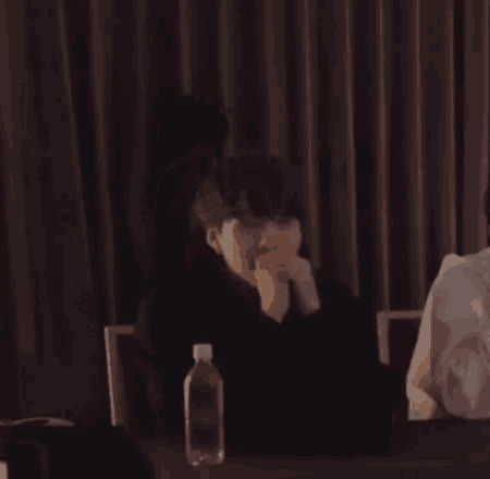 a man is sitting at a table in a dark room with a bottle of water in front of him .
