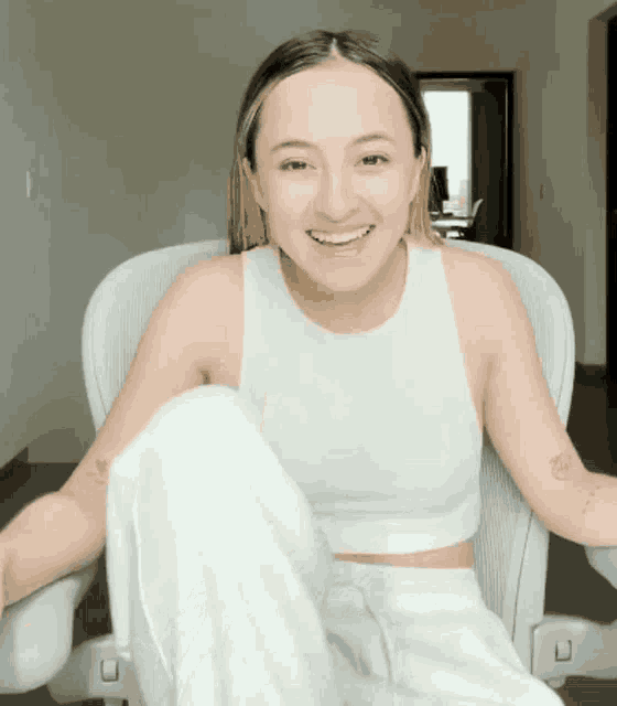 a woman in a white tank top and white pants smiles while sitting in a white chair