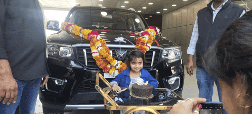 a little girl stands in front of a car with a cake on a cart