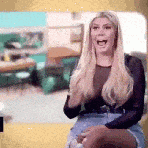 a woman with long blonde hair is sitting in front of a yellow wall and laughing .