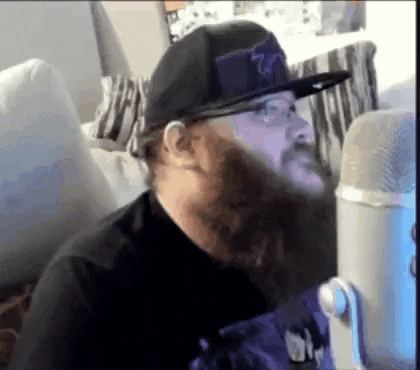 a man with a beard is wearing a hat and glasses while sitting in front of a microphone .