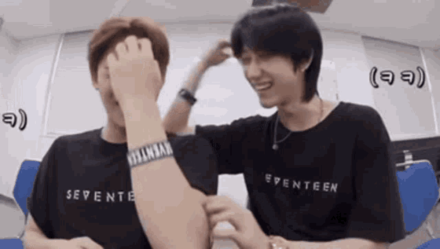 two men are sitting next to each other in a room . one of the men is wearing a seventeen t-shirt .