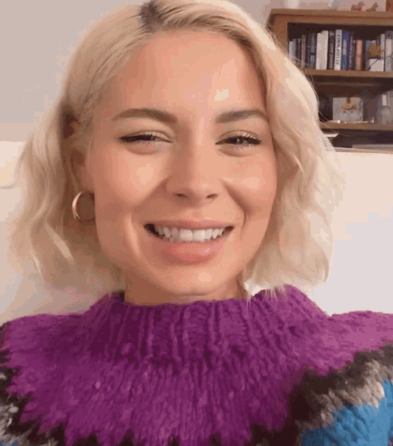 a blonde woman wearing a purple and blue sweater is smiling