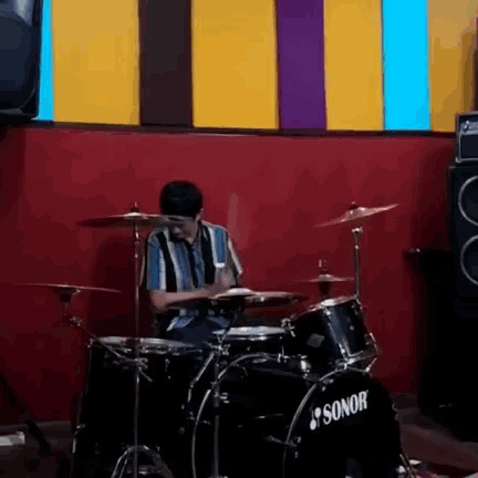 a man is playing drums in a room with a red wall behind him .