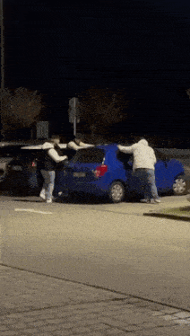a group of men are fighting over a blue car in a parking lot at night .
