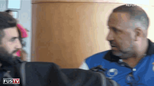 a man in a blue jacket is talking to another man in front of a screen that says fustv