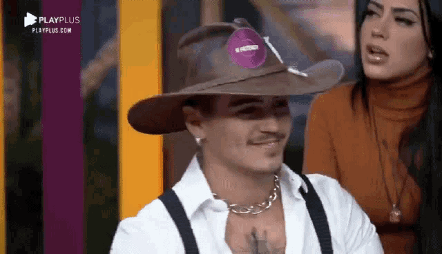 a man wearing a cowboy hat and suspenders is smiling while a woman looks on .