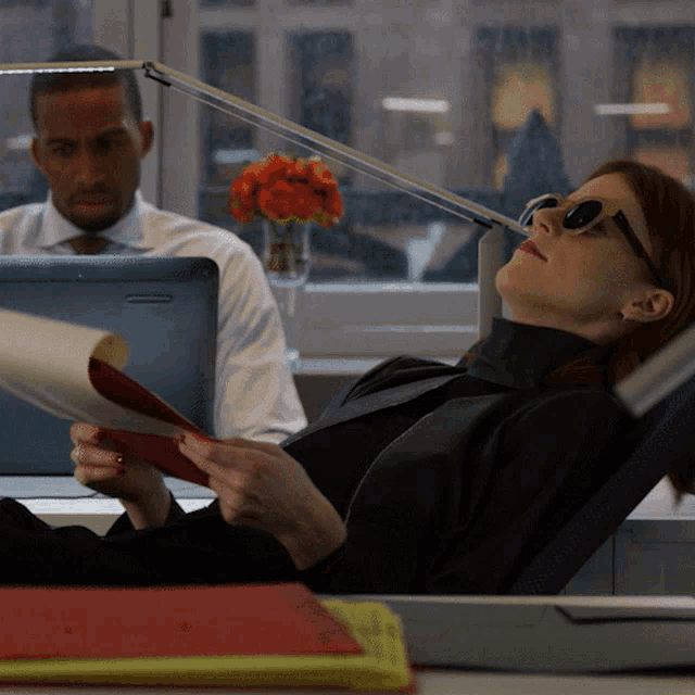 a woman wearing sunglasses is reading a book while a man sits at a desk with a laptop