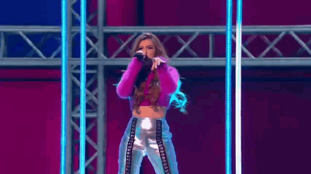 a woman in a pink crop top and silver pants is singing into a microphone on stage .