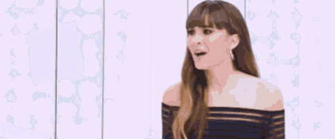 a woman in a black off the shoulder top is standing in front of a white wall with her mouth open .