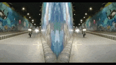 two people riding motorcycles down a street with a mural on the wall