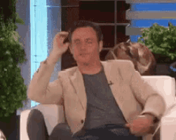 a man in a jacket is sitting in a chair with his legs crossed and scratching his head .