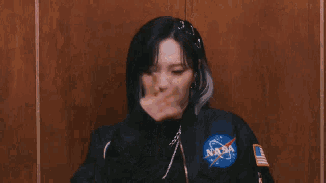 a woman wearing a nasa jacket stands in front of a wooden wall