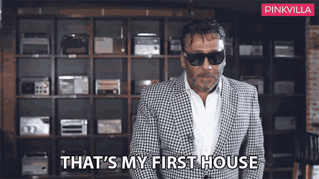 a man in a suit and sunglasses is standing in front of a shelf and says that 's my first house