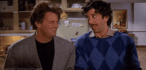 two men are standing next to each other in a kitchen . one of the men is wearing a blue sweater .