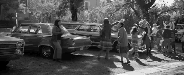 a black and white photo of a group of people dancing in front of a row of cars .