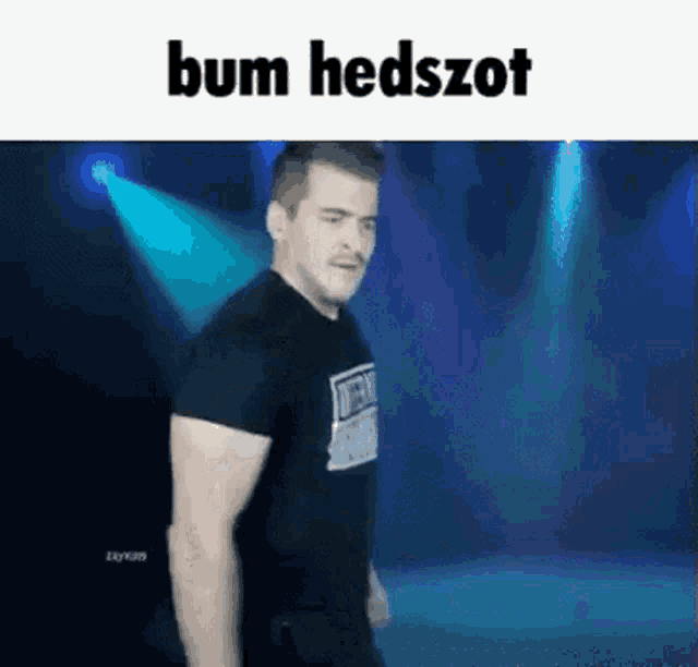 a man in a black shirt is standing on a stage in front of a blue light and says bum hedszot .
