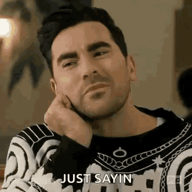 a man in a black and white sweater is sitting at a table with his hand on his chin and says `` just sayin '' .