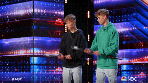 two boys are standing on a stage holding sticks and one is wearing a green sweatshirt .