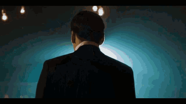 the back of a man in a tuxedo is shown in a dark room with lights hanging from the ceiling .