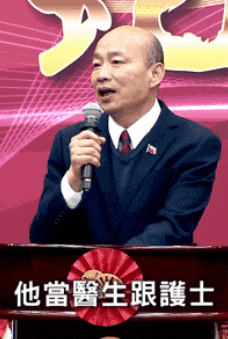 a man in a suit stands at a podium holding a microphone with chinese writing behind him