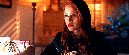 a woman with red hair and red lipstick is sitting at a table with a lamp in the background .