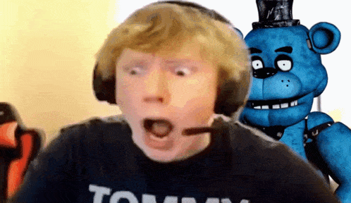 a boy wearing headphones and a shirt that says tommy stands in front of a blue teddy bear