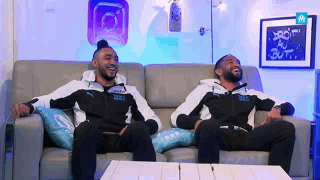 two men are sitting on a couch laughing and one of them has a jacket that says puma