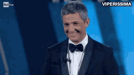 a man in a tuxedo and bow tie is smiling while standing in front of a microphone .