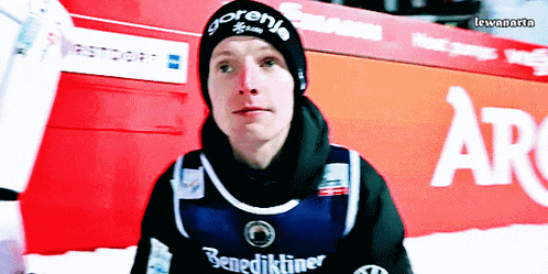 a man wearing a gorenj beanie stands in front of a red sign