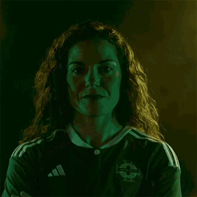 a woman with curly hair wearing a green adidas jersey looks at the camera