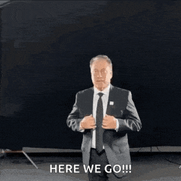 a man in a suit and tie is standing in front of a black curtain and says `` here we go '' .