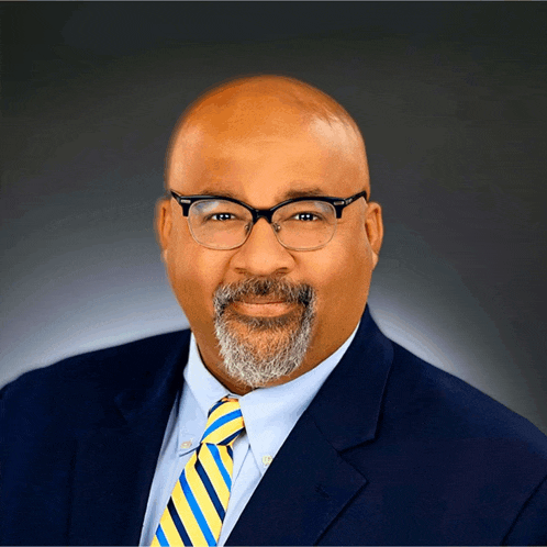 a man with glasses and a beard wears a blue suit and yellow and blue striped tie