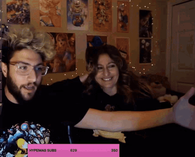 a man and a woman are sitting in front of a wall with posters on it and a pink box that says hypemas subs