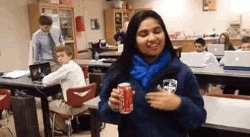 a girl in a blue jacket is holding a can of soda in a classroom .
