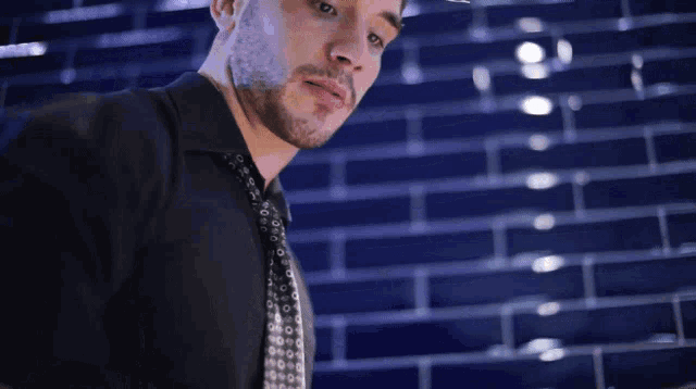 a man in a black jacket and tie is standing in front of a blue brick wall