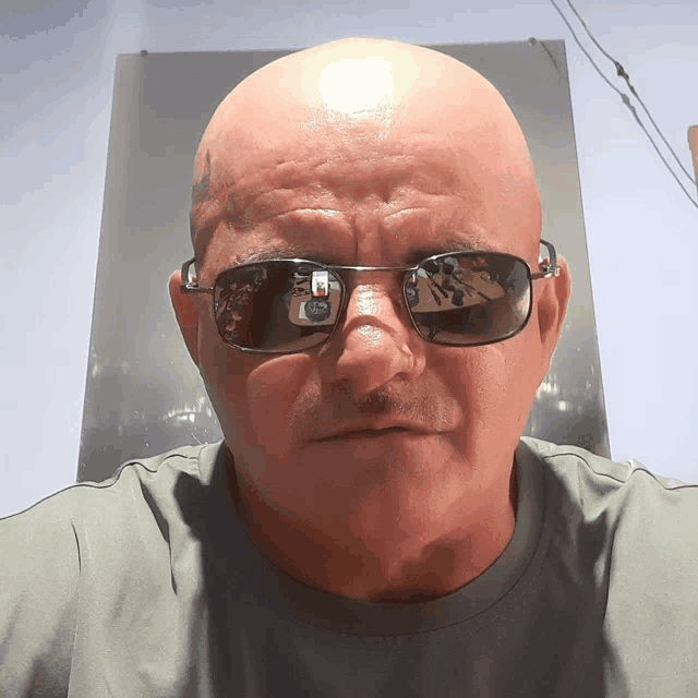 a bald man wearing sunglasses looks at the camera with a reflection of himself in the sunglasses