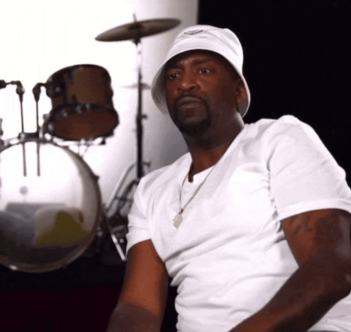 a man wearing a white hat and a white shirt is sitting in front of drums