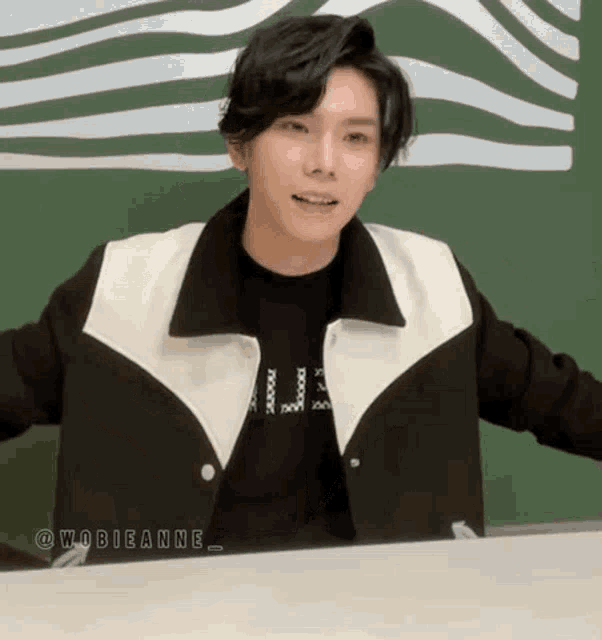 a young man is sitting at a table wearing a black and white jacket and a black shirt .