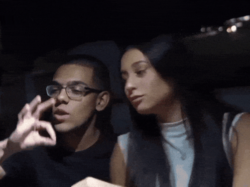 a man with glasses is smoking a cigarette while a woman holds his hand over his mouth