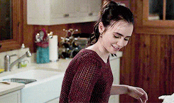 a woman in a red sweater is standing in a kitchen next to a sink and smiling .
