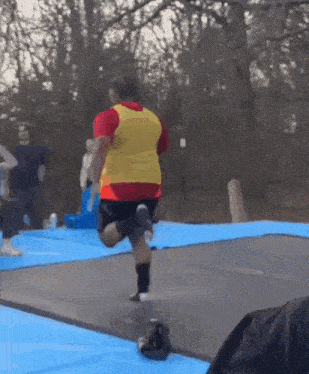 a man in a red shirt and yellow vest is jumping on a blue mat on a trampoline .