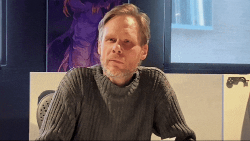 a man in a grey sweater sits in front of a purple poster