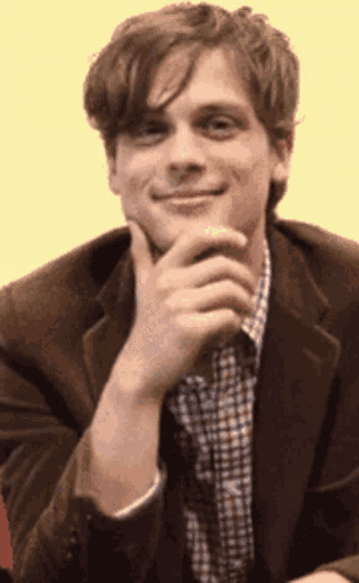 a man in a suit and plaid shirt holds his hand to his chin and smiles