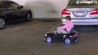 Criança de gravata e óculos escuros dirigindo um pequeno carro elétrico. 