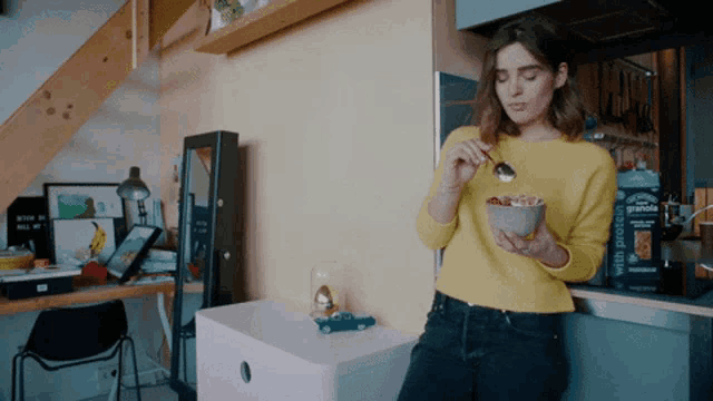 a woman in a yellow sweater is eating a bowl of granola with a spoon