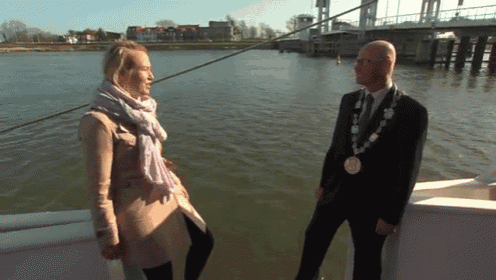 a man with a chain around his neck talks to a woman in front of a body of water